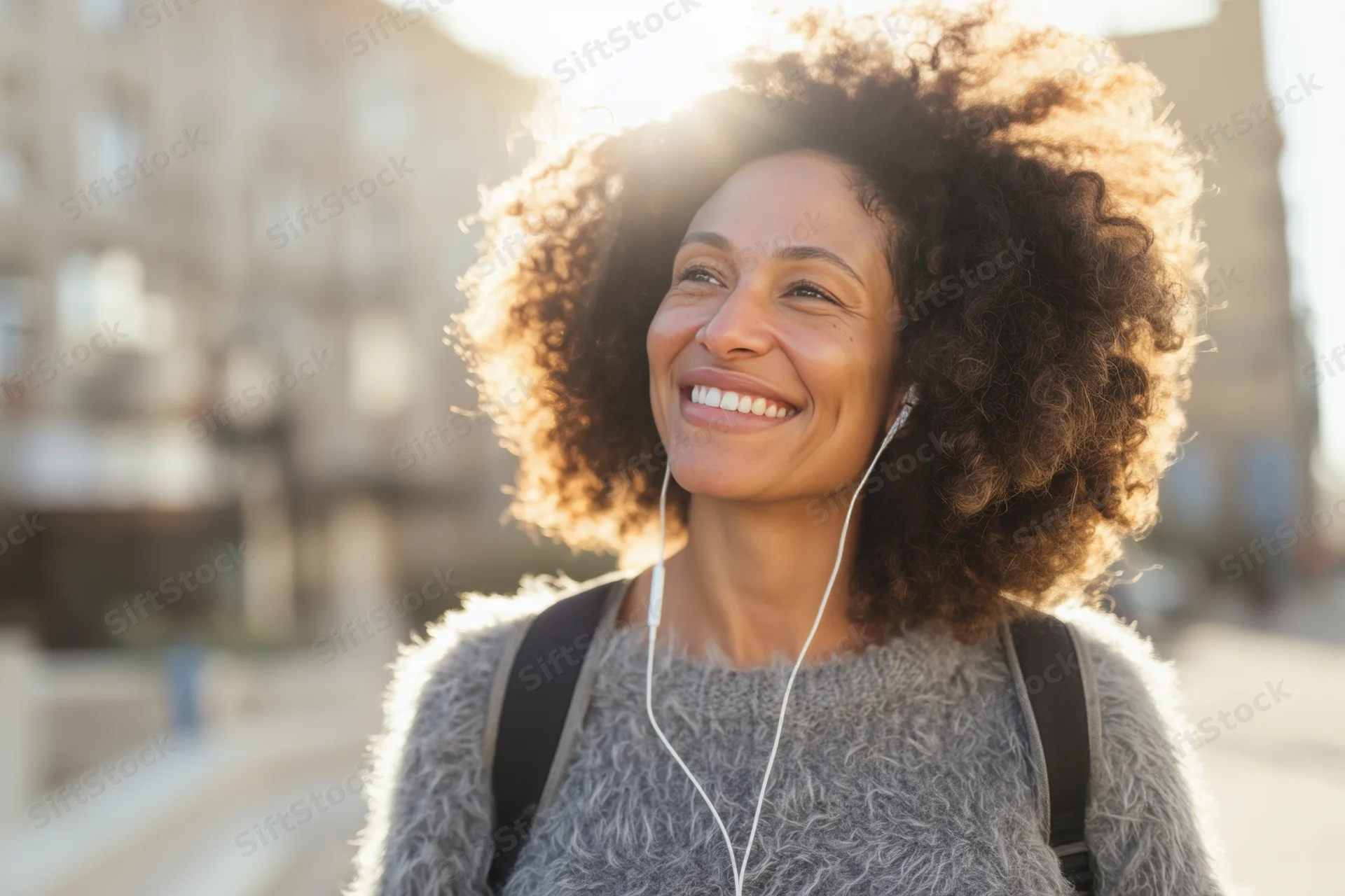 Woman with voluminous curly afro smiling outdoors in sunlight wearing earphones and backpack