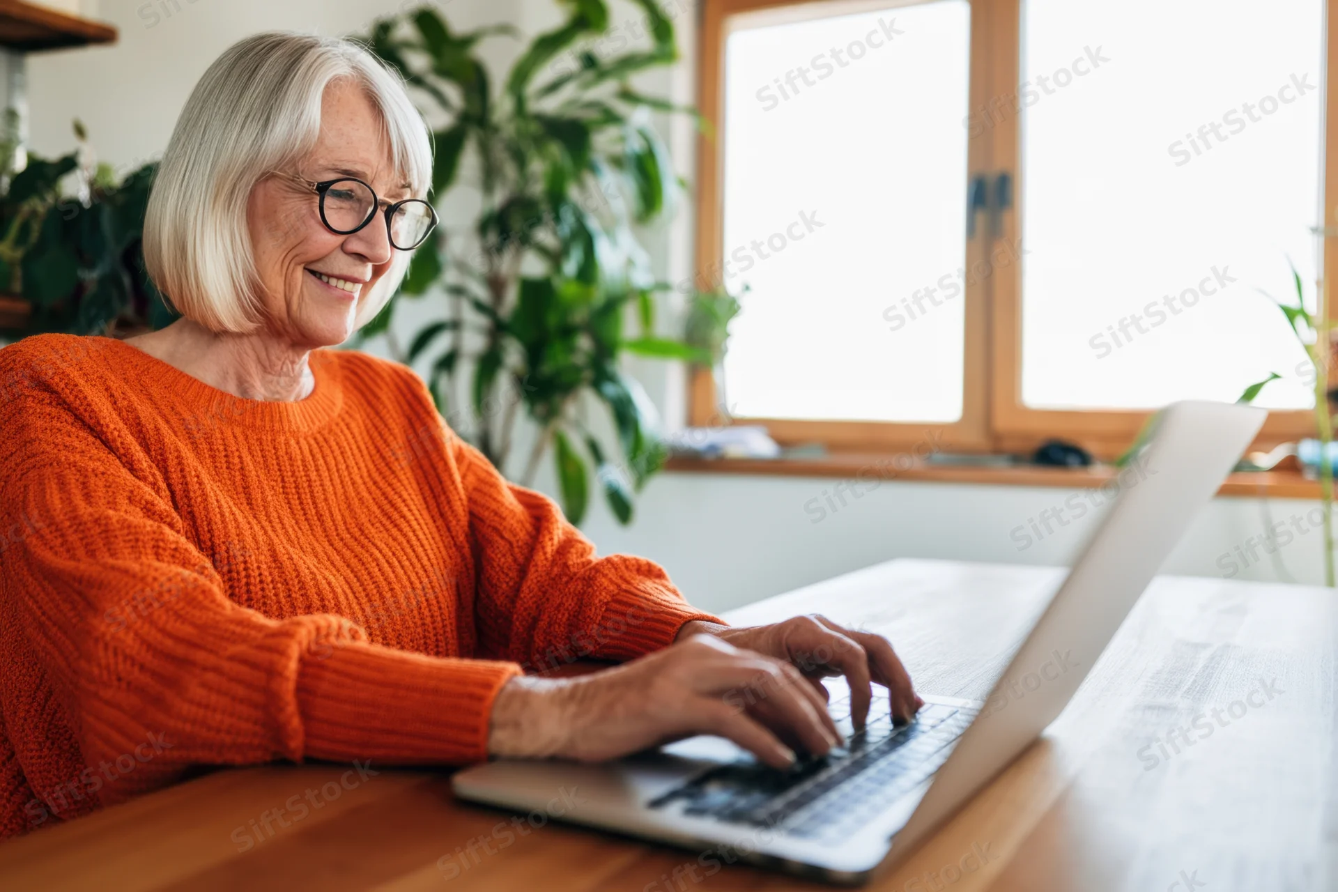 Senior woman typing on laptop at wooden desk by window in home office