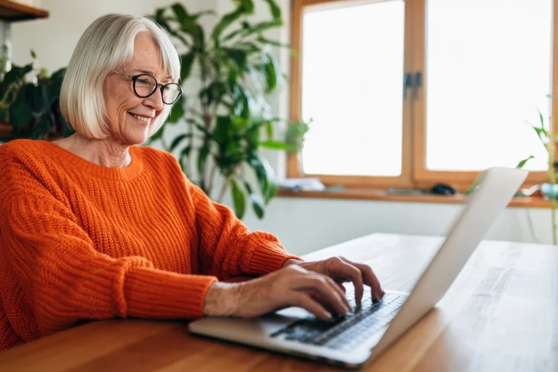 Senior woman typing on laptop at wooden desk by window in home office
