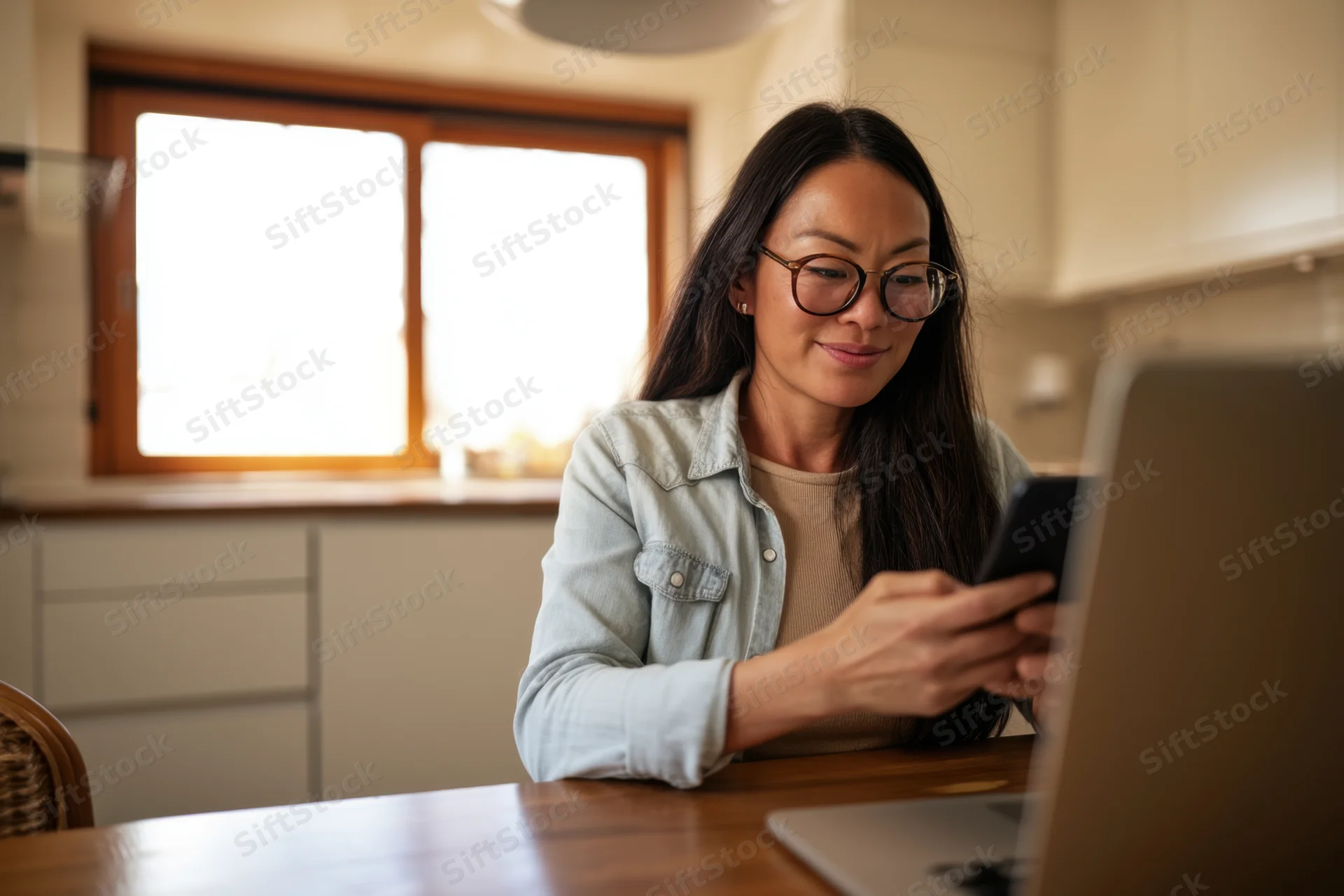 Woman using smartphone while sitting at wooden table in kitchen with laptop open in front of her