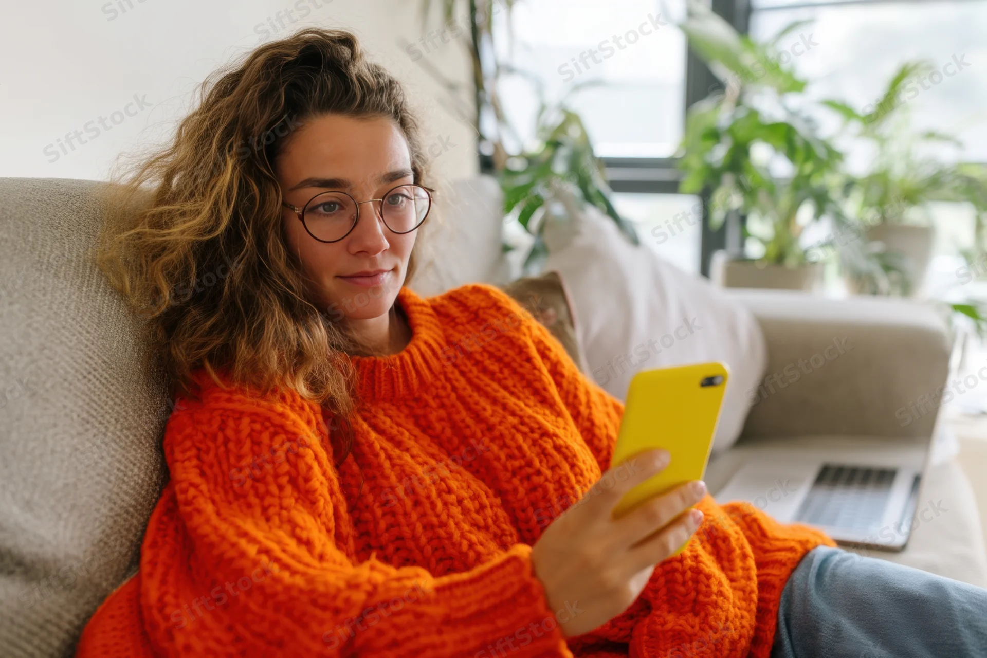 Woman in orange sweater relaxing on couch with smartphone in hand