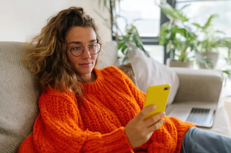 Woman in orange sweater relaxing on couch with smartphone in hand