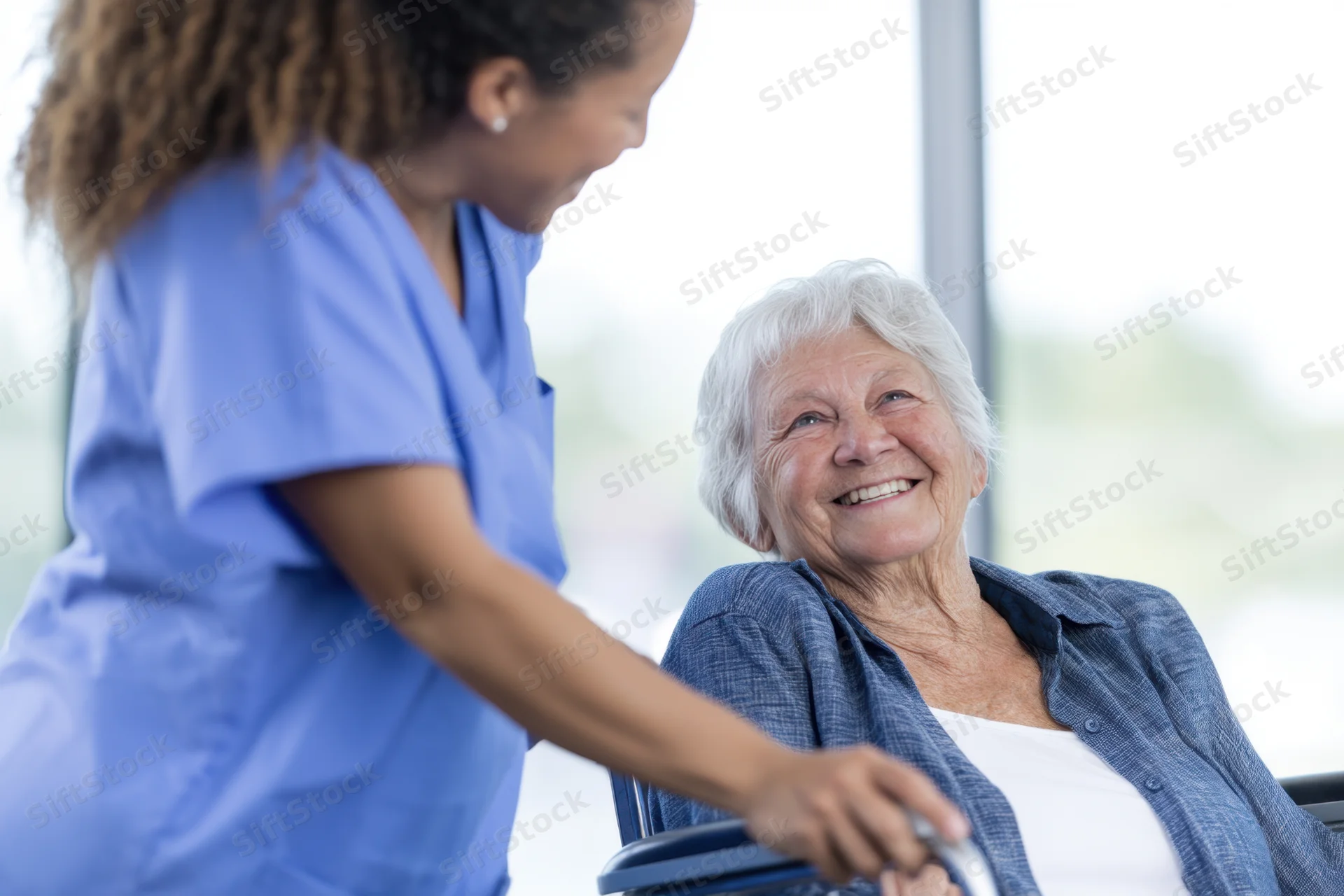 Elderly woman in wheelchair smiling as caregiver in blue scrubs assists her indoors