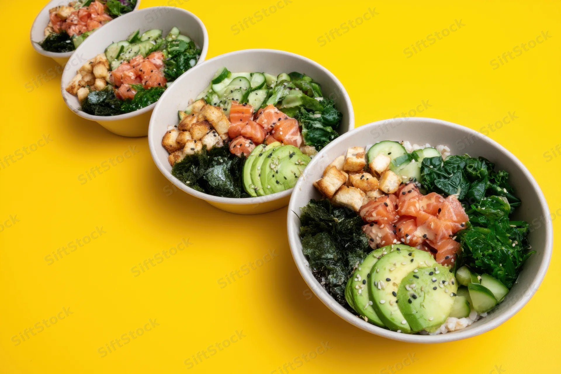 Five poke bowls with salmon, avocado, cucumber, seaweed, tofu and sesame arranged in a row on a yellow background
