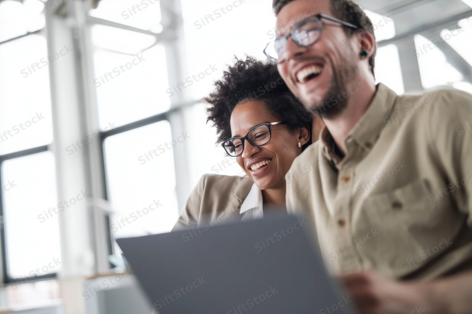 Two coworkers laughing while looking at a laptop in a bright modern office