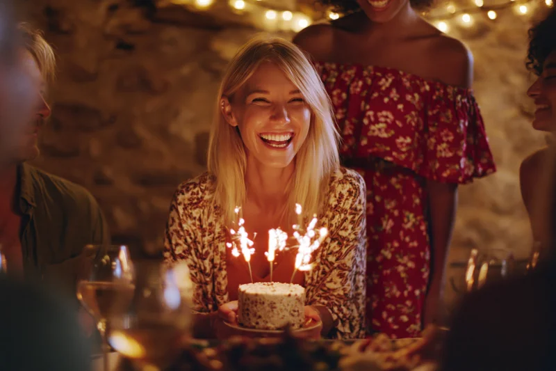 Blonde woman laughing while holding a small birthday cake with sparklers at a dinner party with friends