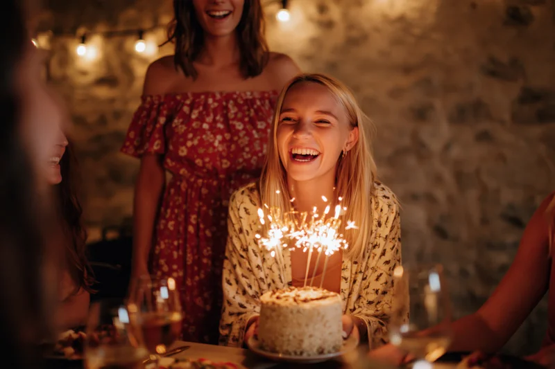 Blonde woman laughing while holding a small birthday cake with sparklers at a dinner party