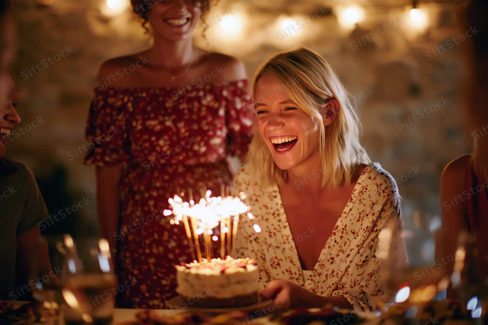 Blonde woman laughing while holding a small birthday cake with sparklers at an indoor evening party