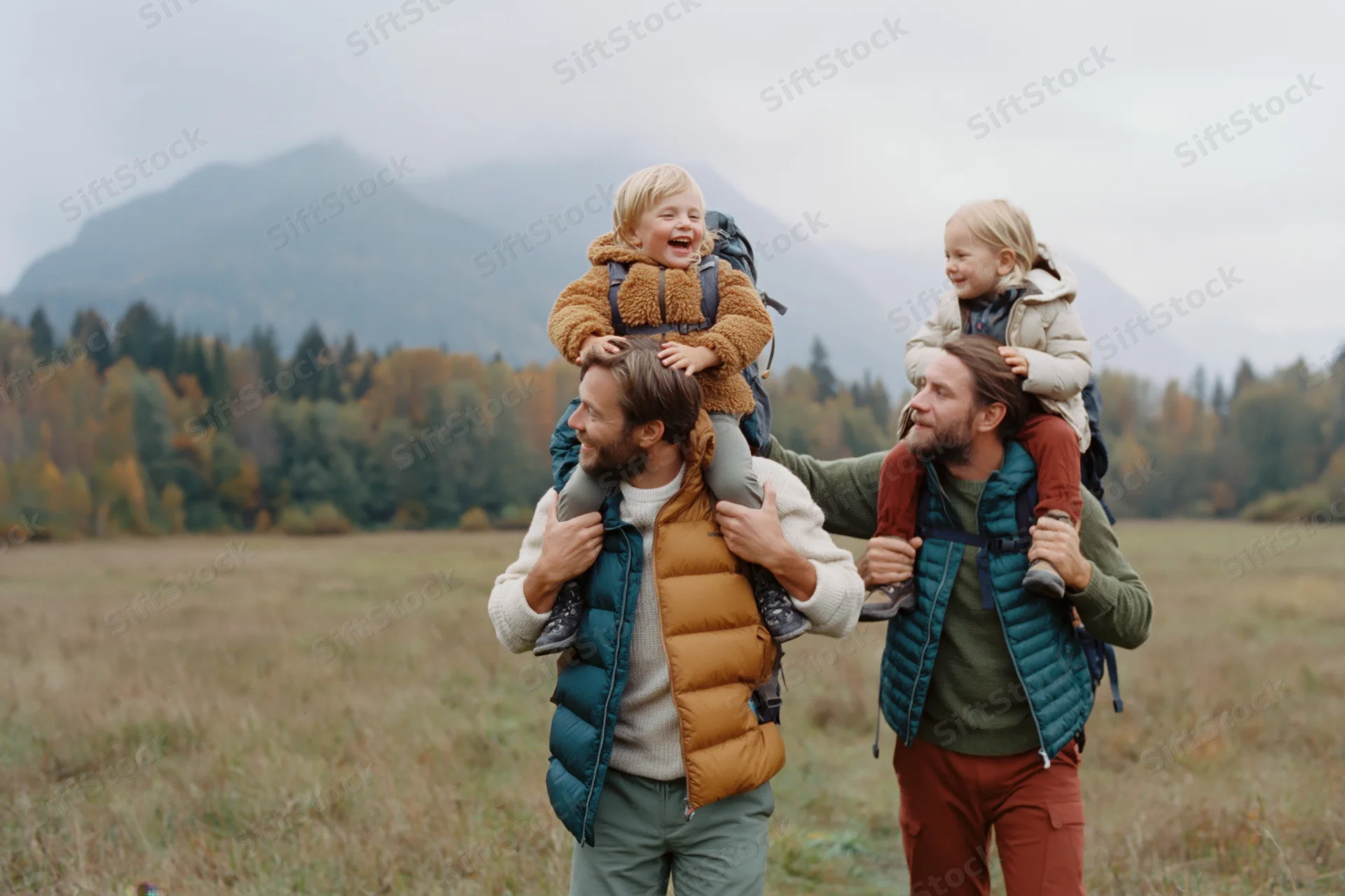 Two fathers carrying their young children on shoulders while hiking through an autumn field with mountains in the background