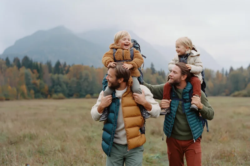 Two fathers carrying their young children on shoulders while hiking through an autumn field with mountains in the background