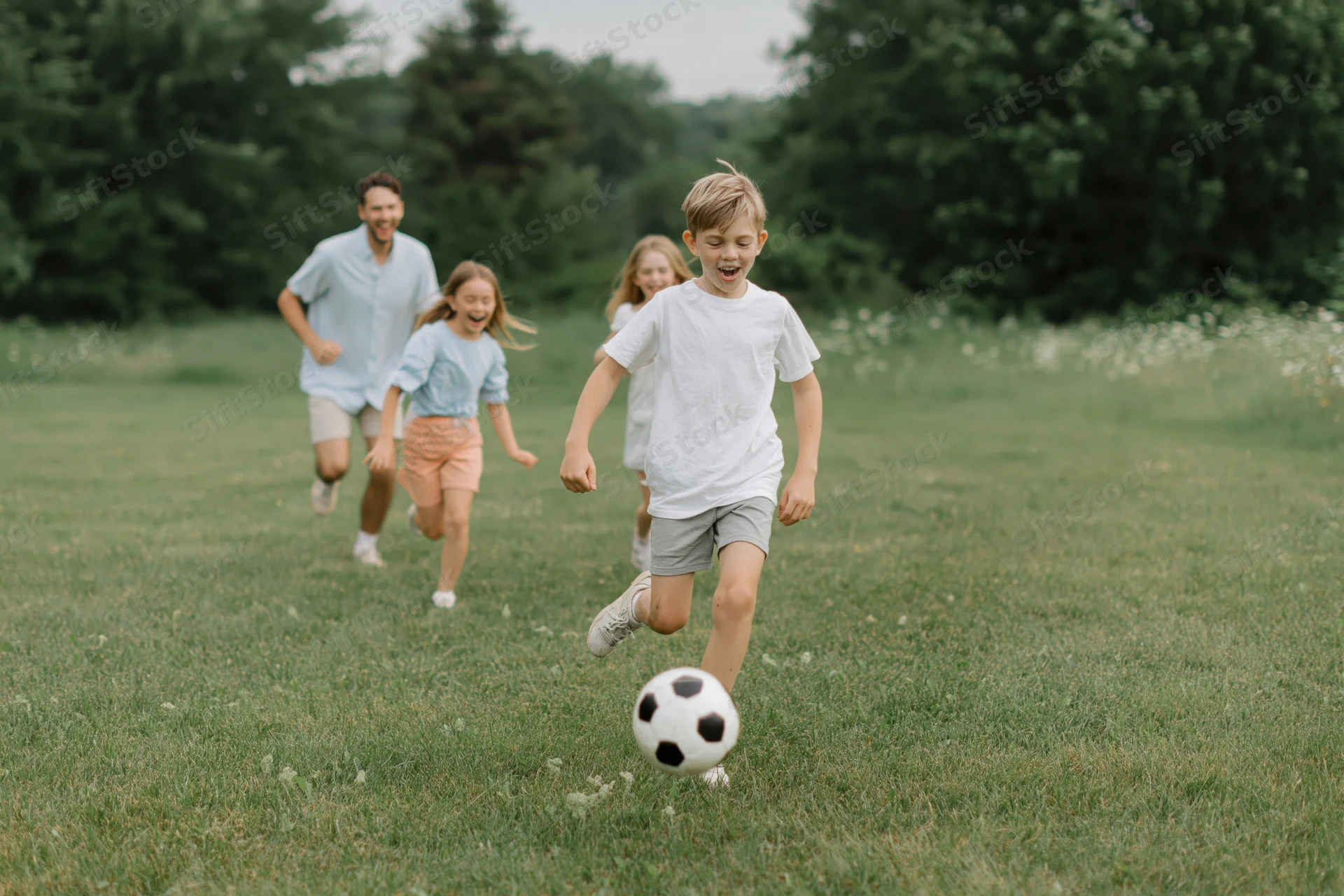 Boy kicking soccer ball in grassy park with family running behind