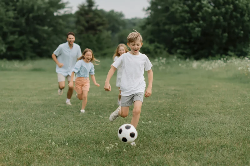 Boy kicking soccer ball in grassy park with family running behind