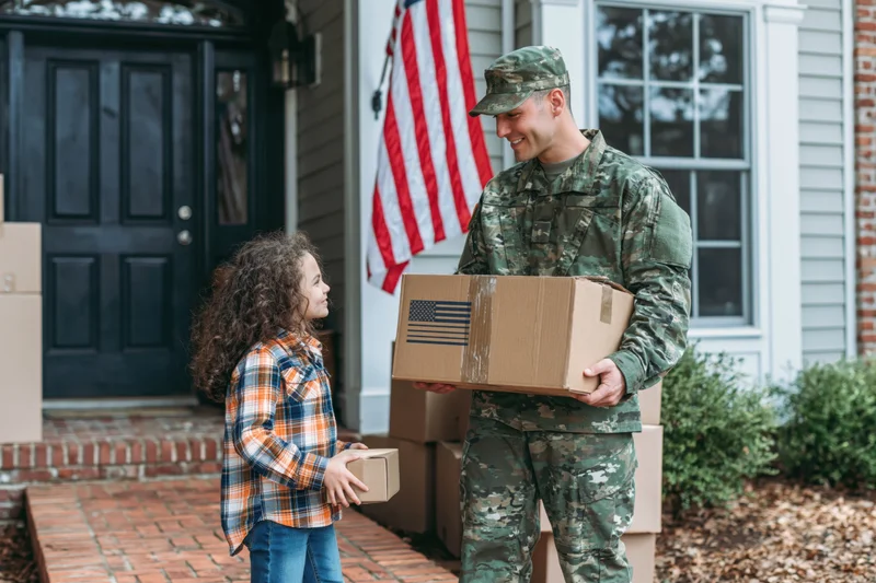 Soldier in camouflage uniform holding box on porch with daughter holding small box, american flag in background