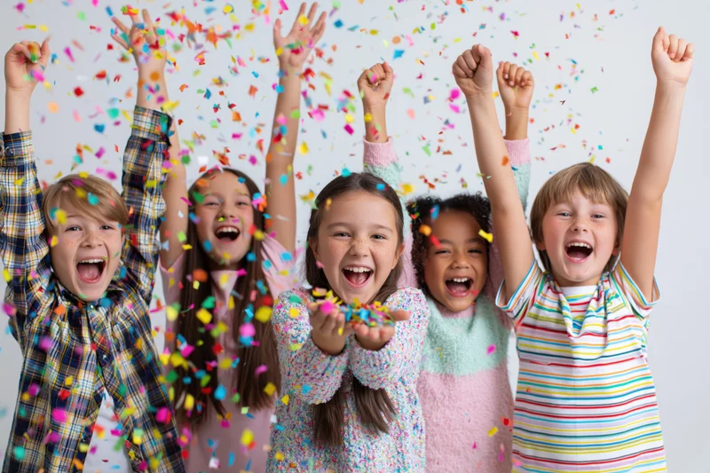 Five children cheering with arms raised amidst falling confetti in a studio setting