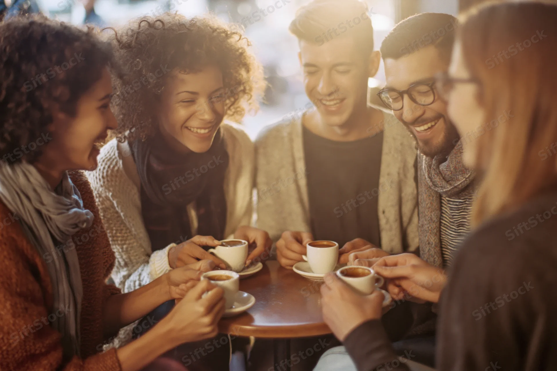 Five friends laughing together around a small wooden table in a cafe holding coffee cups
