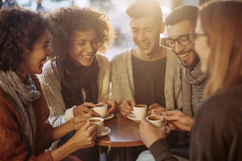 Five friends laughing together around a small wooden table in a cafe holding coffee cups