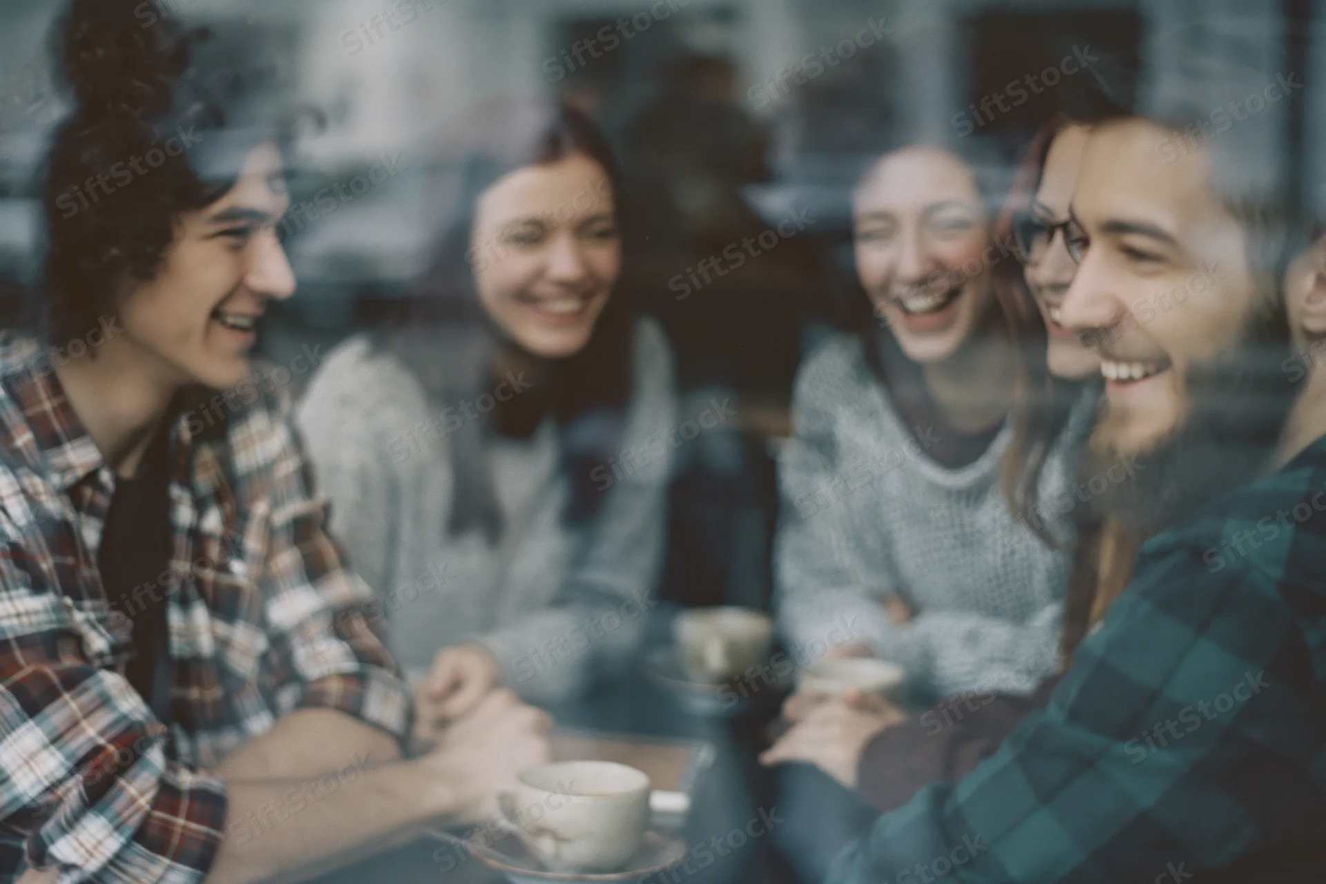 Five young friends laughing together around a cafe table viewed through a window