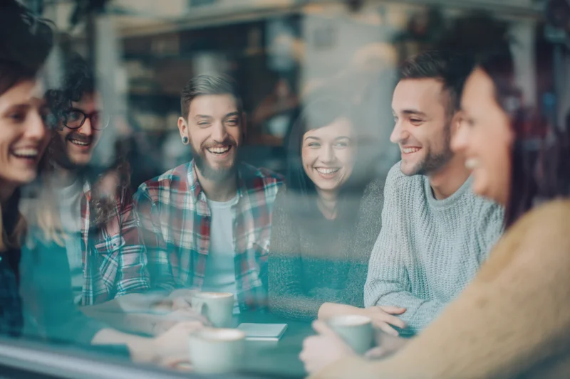 Five young friends laughing together around a cafe table viewed through a window