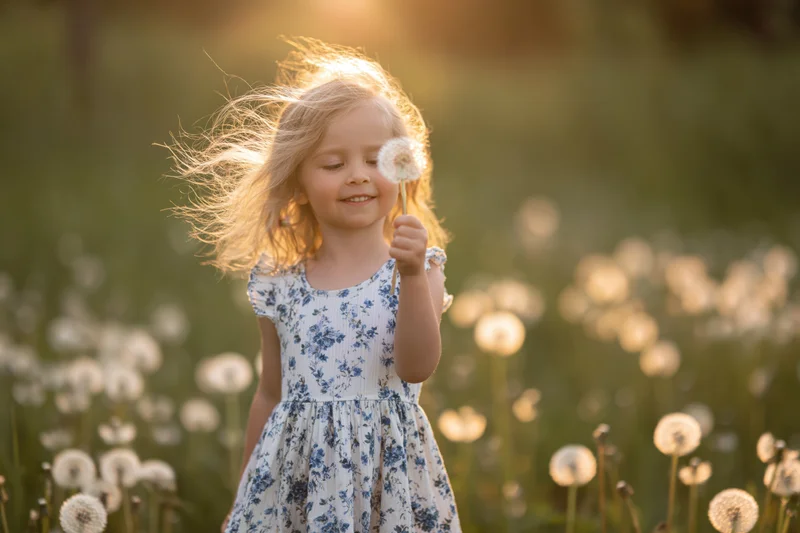 Young blonde girl smiling while holding a dandelion in a sunlit meadow