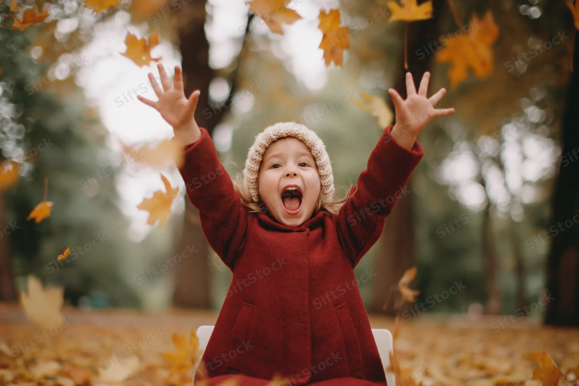Young girl sitting on ground in autumn park with arms raised amid falling leaves