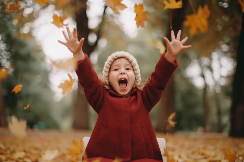 Young girl sitting on ground in autumn park with arms raised amid falling leaves