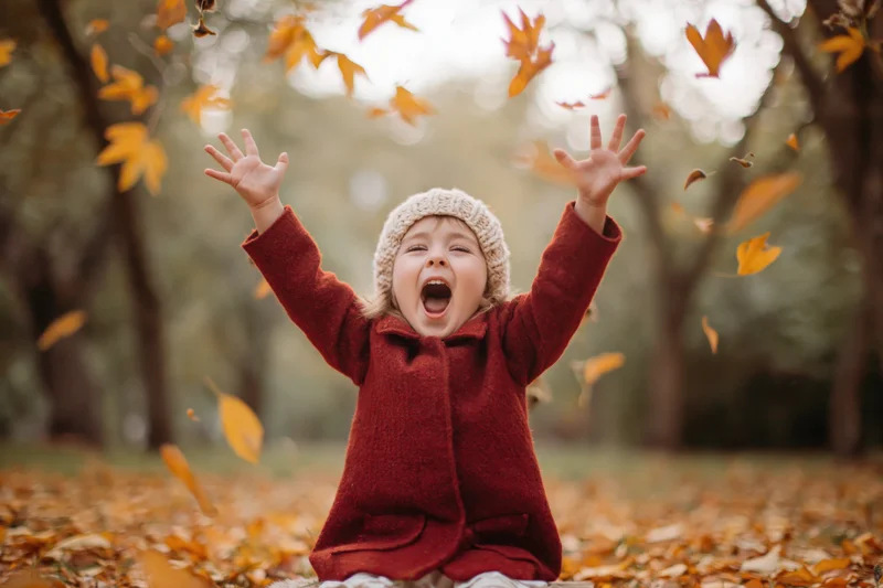 Child sitting on ground in autumn park with arms raised and leaves falling around