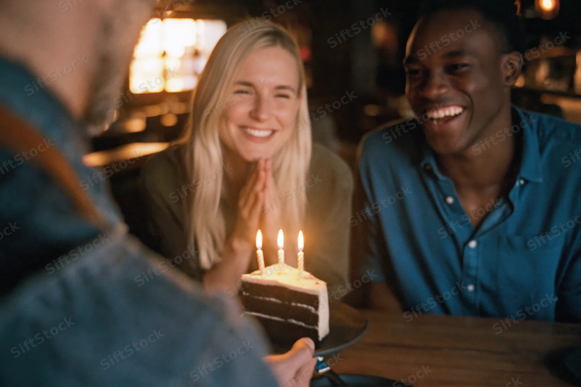 Friends smiling at a slice of birthday cake with lit candles on a wooden table in a restaurant