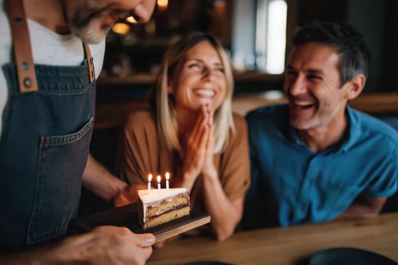 Waiter presenting slice of cake with three lit candles to smiling couple in restaurant