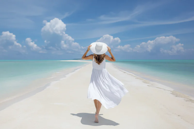 Woman walking barefoot along a narrow sandbank toward turquoise ocean under blue sky with clouds