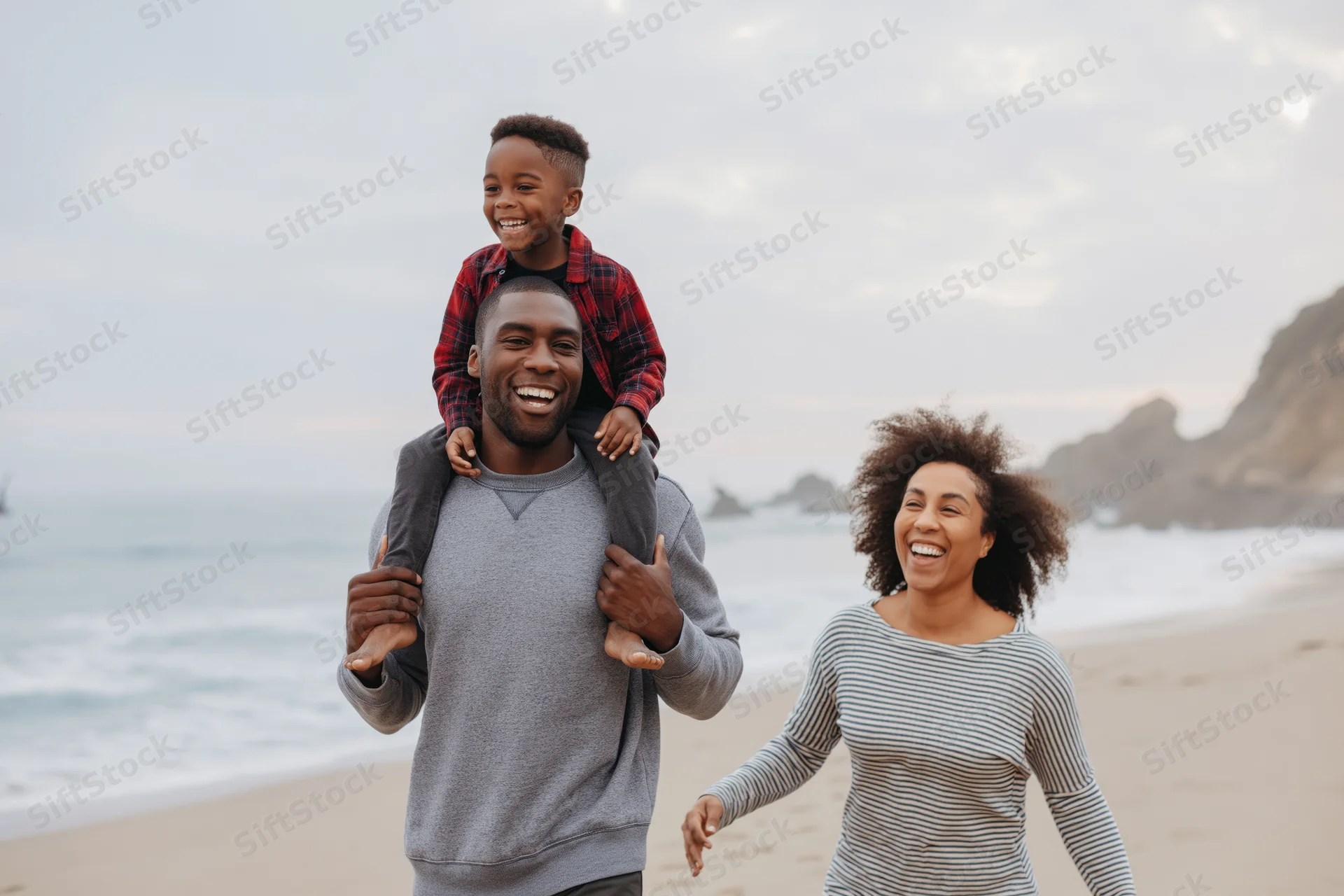 Young black family walking on sandy beach with father carrying child on shoulders and mother laughing beside them