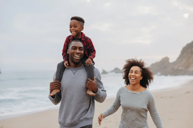 Young black family walking on sandy beach with father carrying child on shoulders and mother laughing beside them