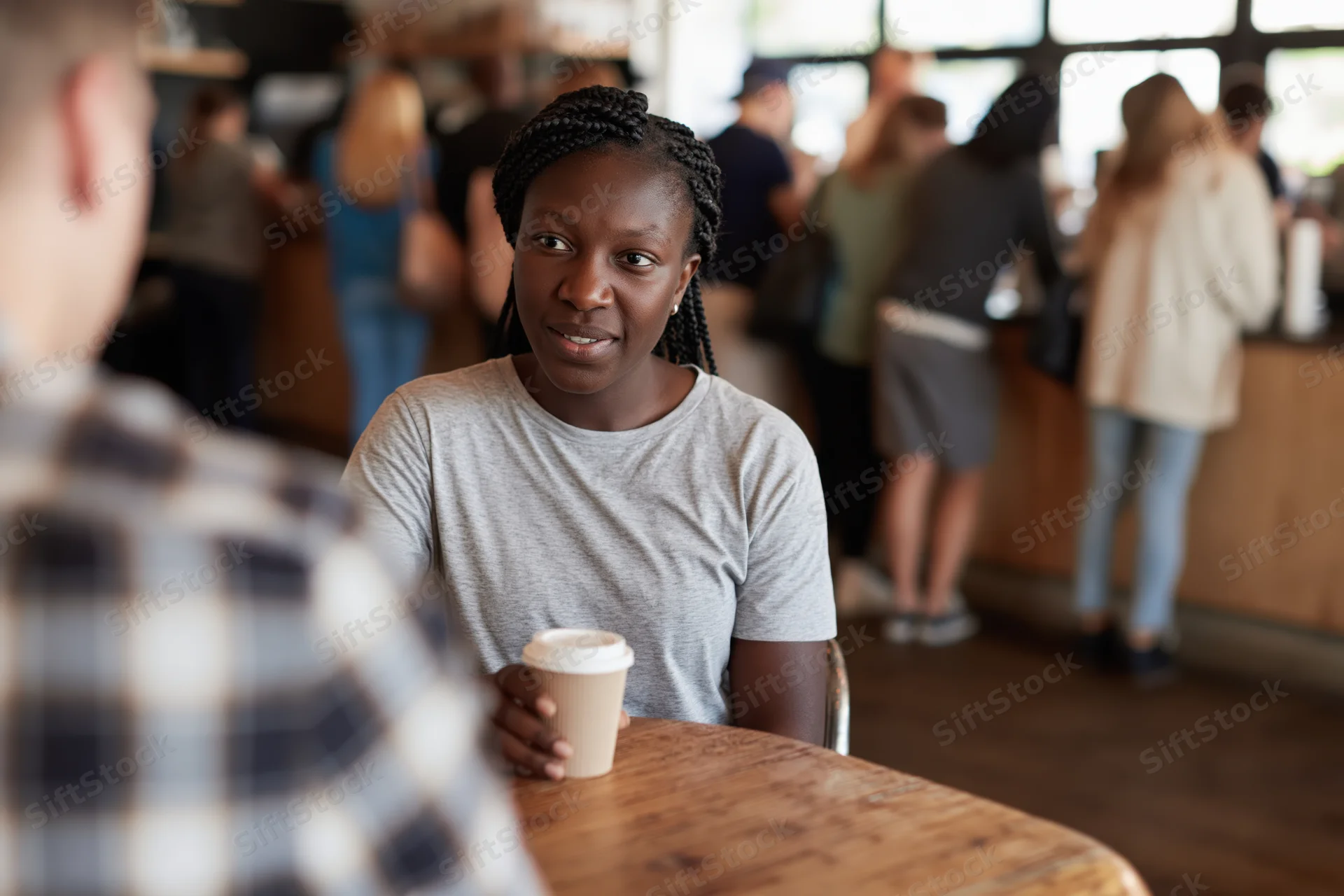 Young black woman with braided hair sitting at wooden table in cafe holding coffee cup, smiling during conversation