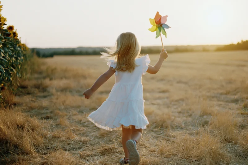 Young blonde girl running through a sunlit field holding a pinwheel