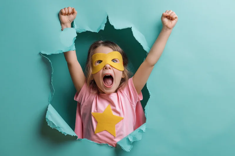 Young child in superhero costume bursting through torn paper with arms raised and mouth open