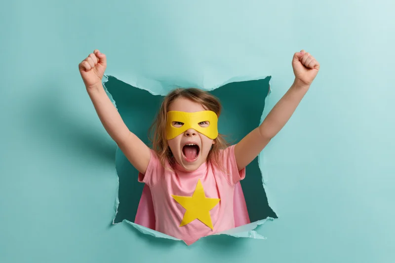 Young girl in superhero costume bursting through paper hole with arms raised and shouting