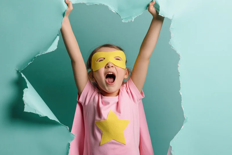 Young girl in homemade superhero costume with yellow mask and pink cape tearing through paper wall