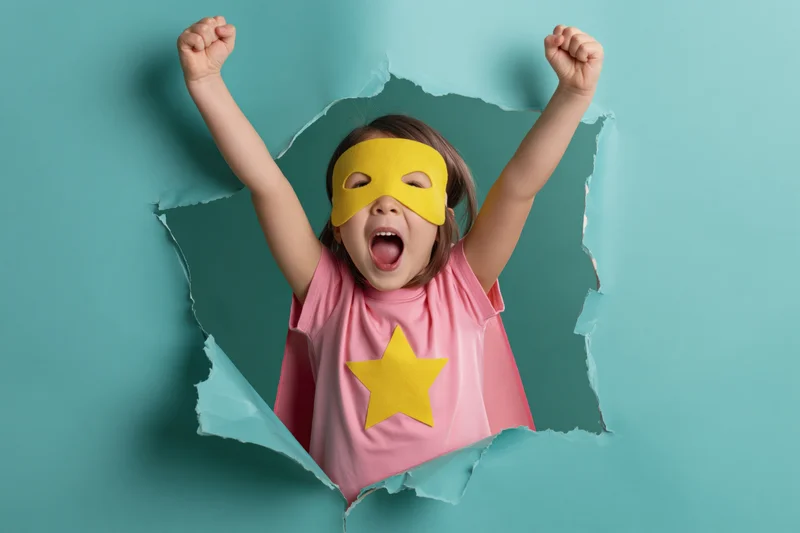 Young girl in superhero costume bursting through paper with arms raised and shouting