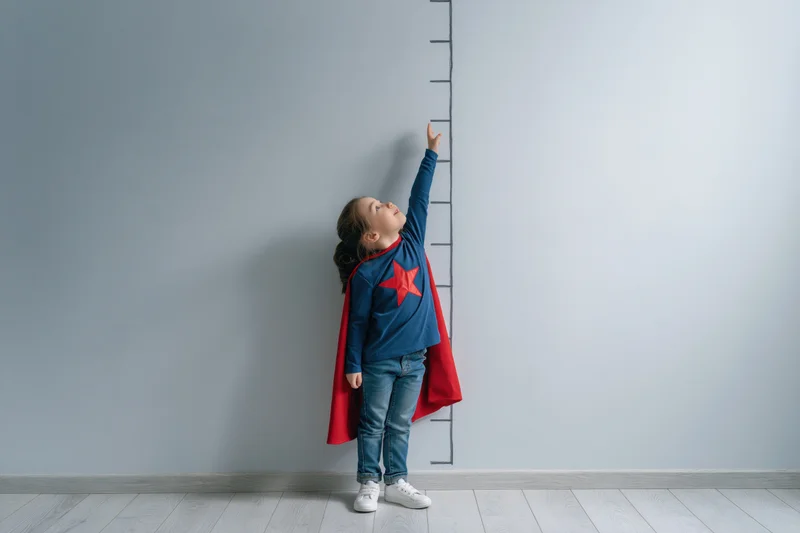 Young girl in superhero costume reaching upward to measure height against wall ruler