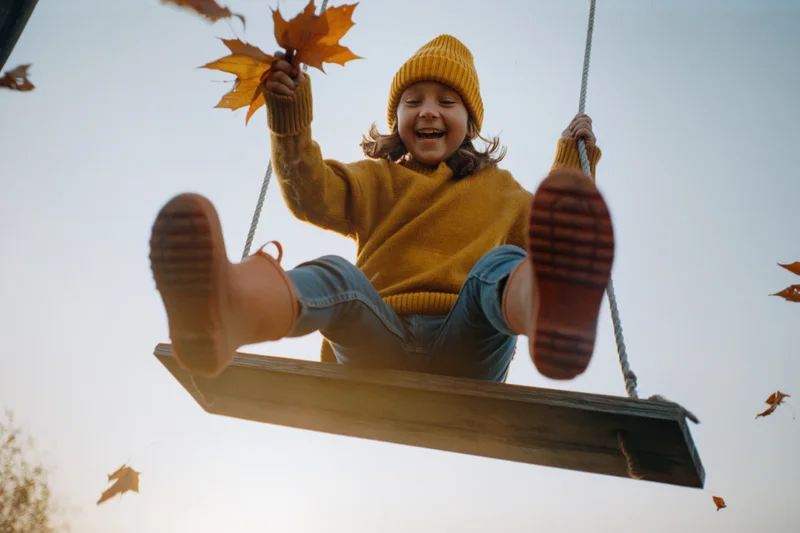 Young girl laughing on wooden swing with autumn leaves swirling around her