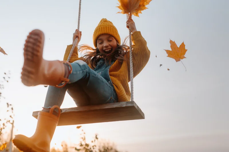 Young girl laughing on wooden swing holding autumn leaves outdoors