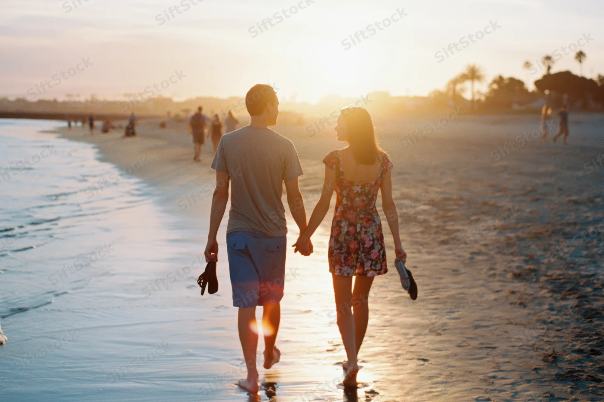 Young couple holding hands walking barefoot along wet beach at sunset