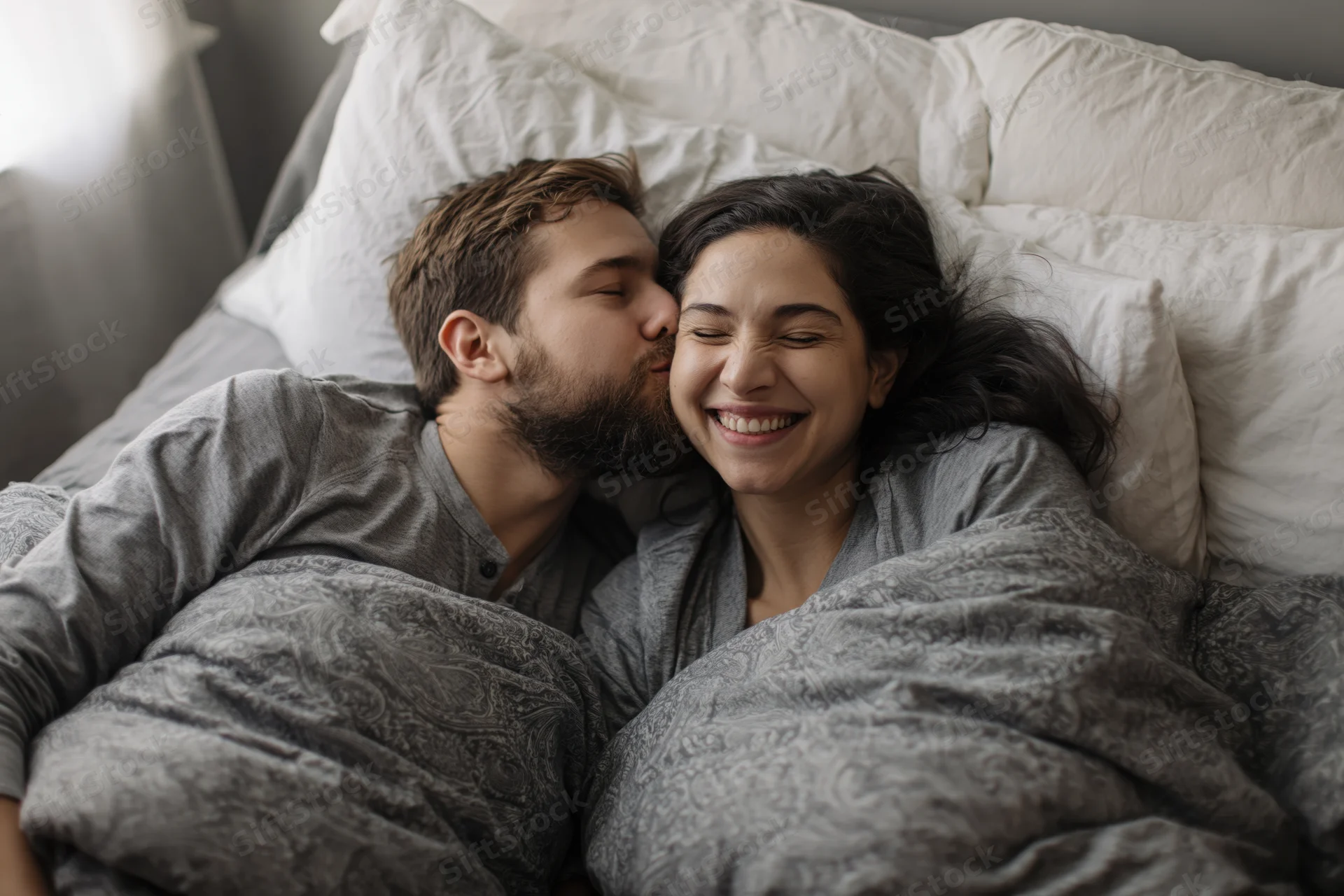 Young couple cuddling in bed with man kissing woman's cheek, both smiling under gray blanket