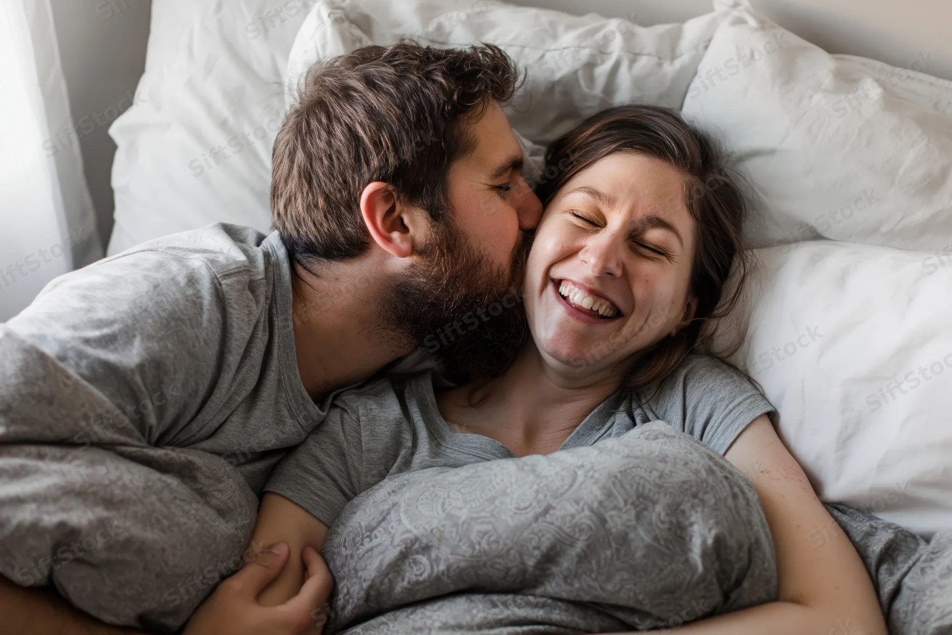 Young couple cuddling in bed with man kissing woman's cheek, both smiling, covered in gray blanket, white pillows behind them