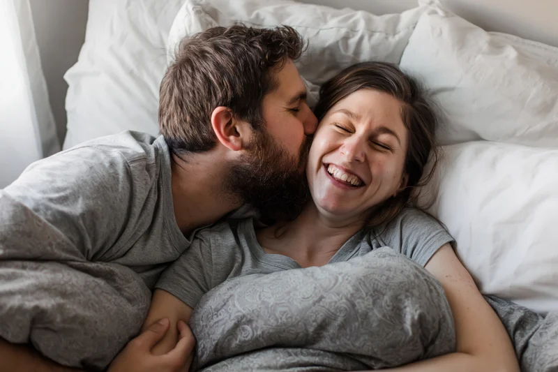 Young couple cuddling in bed with man kissing woman's cheek, both smiling, covered in gray blanket, white pillows behind them