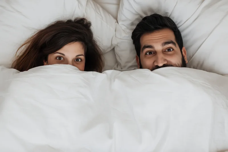 Young couple peeking playfully from under white blanket in bed