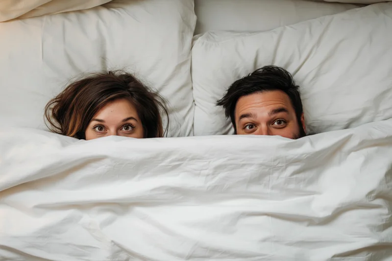 Young couple peeking playfully from under white blanket in bed