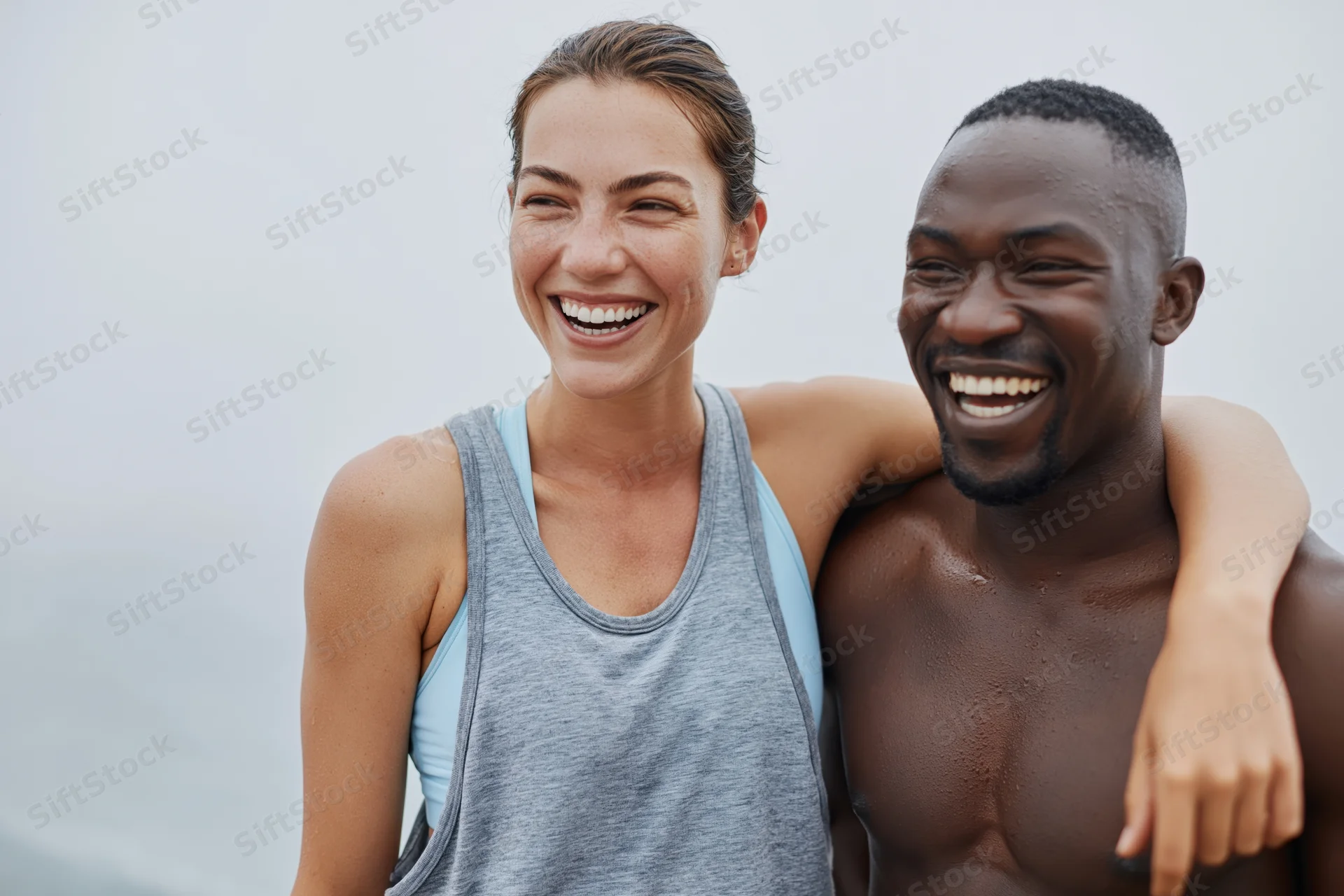 Young interracial couple laughing together outdoors near the ocean, woman in gray tank top, man shirtless, arms around each other