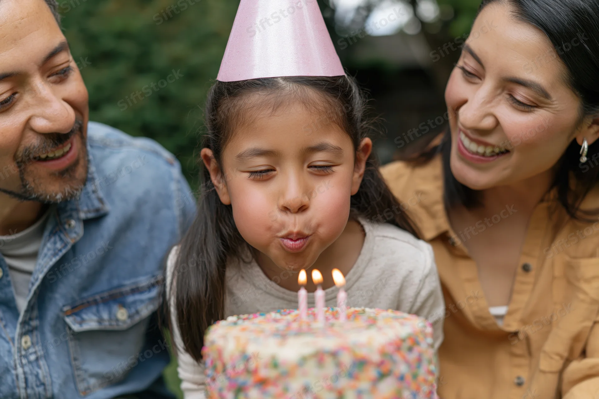 Young mixed-race girl blowing out candles on birthday cake with parents smiling beside her in garden