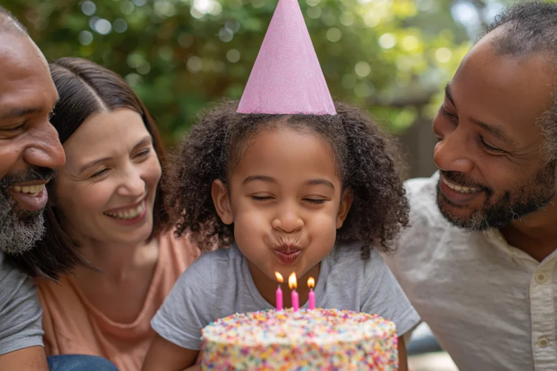 Young mixed-race girl blowing out candles on birthday cake surrounded by family in garden