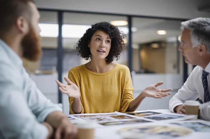 Woman in yellow top gesturing during office meeting with two men at table with documents and coffee cups